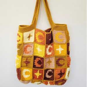 May include: A handmade crochet tote bag in shades of yellow, brown, and cream. The bag features a patchwork design with geometric shapes and symbols. The bag has two long handles and is hanging against a white wall.