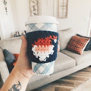 May include: A blue and white crocheted coffee cup cozy with a red, white, and pink design. The cozy is on a white and blue checkered coffee cup.