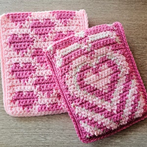 Puede incluir: Dos agarraderas de olla de crochet rosa y blanco. La agarradera m&aacute;s grande est&aacute; en la parte inferior y la m&aacute;s peque&ntilde;a est&aacute; encima de la m&aacute;s grande.