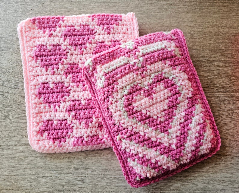 Puede incluir: Dos agarraderas de olla de crochet rosa y blanco. La agarradera m&aacute;s grande est&aacute; en la parte inferior y la m&aacute;s peque&ntilde;a est&aacute; encima de la m&aacute;s grande.
