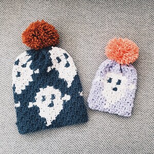 May include: Two crocheted hats with ghost designs. The larger hat is blue with a white ghost design and an orange pom-pom. The smaller hat is purple with a white ghost design and a pink pom-pom.