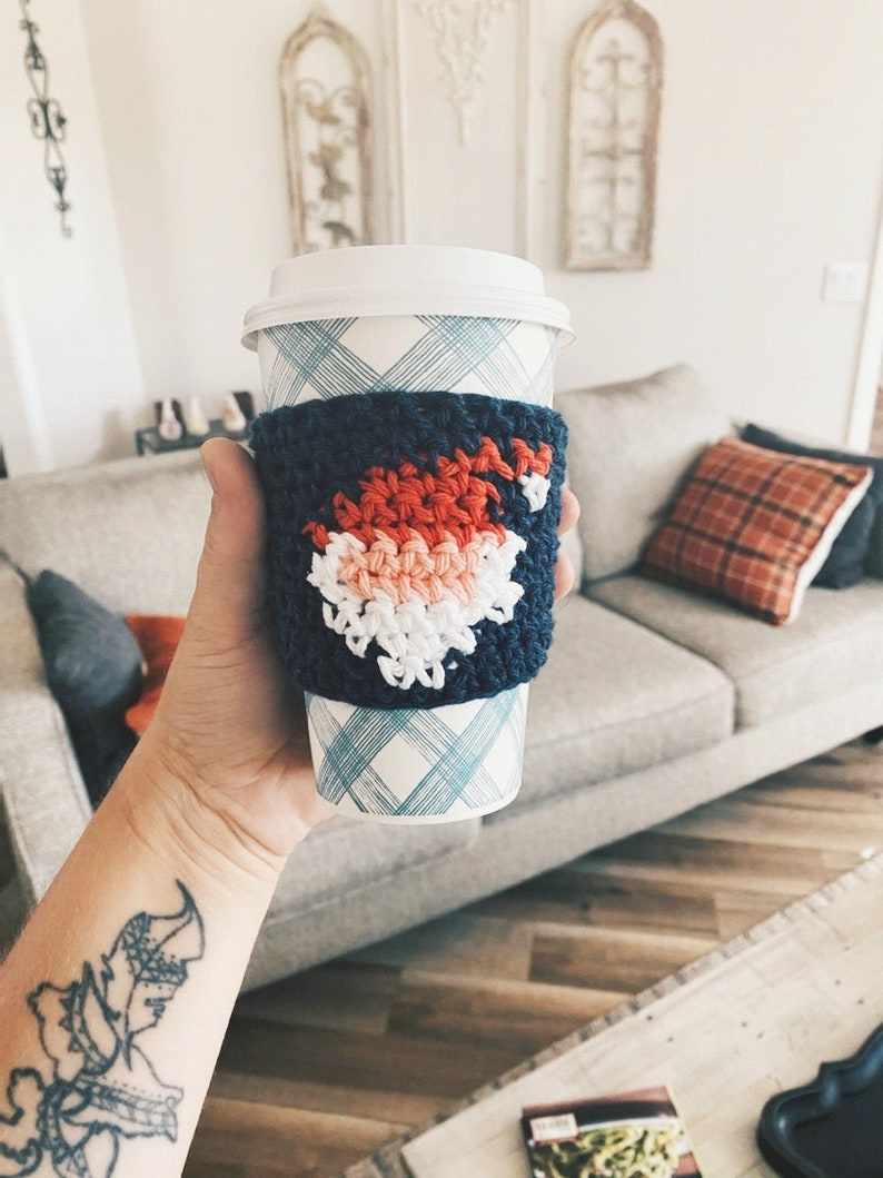 May include: A blue and white crocheted coffee cup cozy with a red, white, and pink design. The cozy is on a white and blue checkered coffee cup.