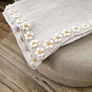 May include: A gray crocheted blanket with a white and yellow daisy border. The blanket is folded and placed on a gray textured surface.