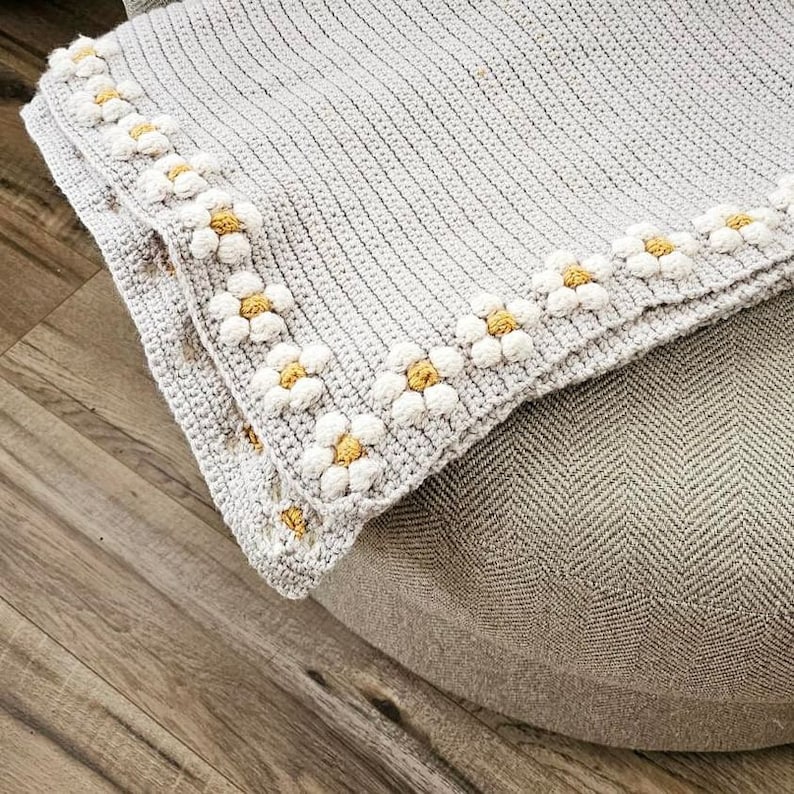 May include: A gray crocheted blanket with a white and yellow daisy border. The blanket is folded and placed on a gray textured surface.