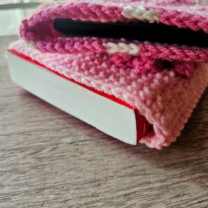 Puede incluir: Una funda de tel&eacute;fono de crochet rosa y blanca con una franja blanca, que cubre un libro con una encuadernaci&oacute;n roja.