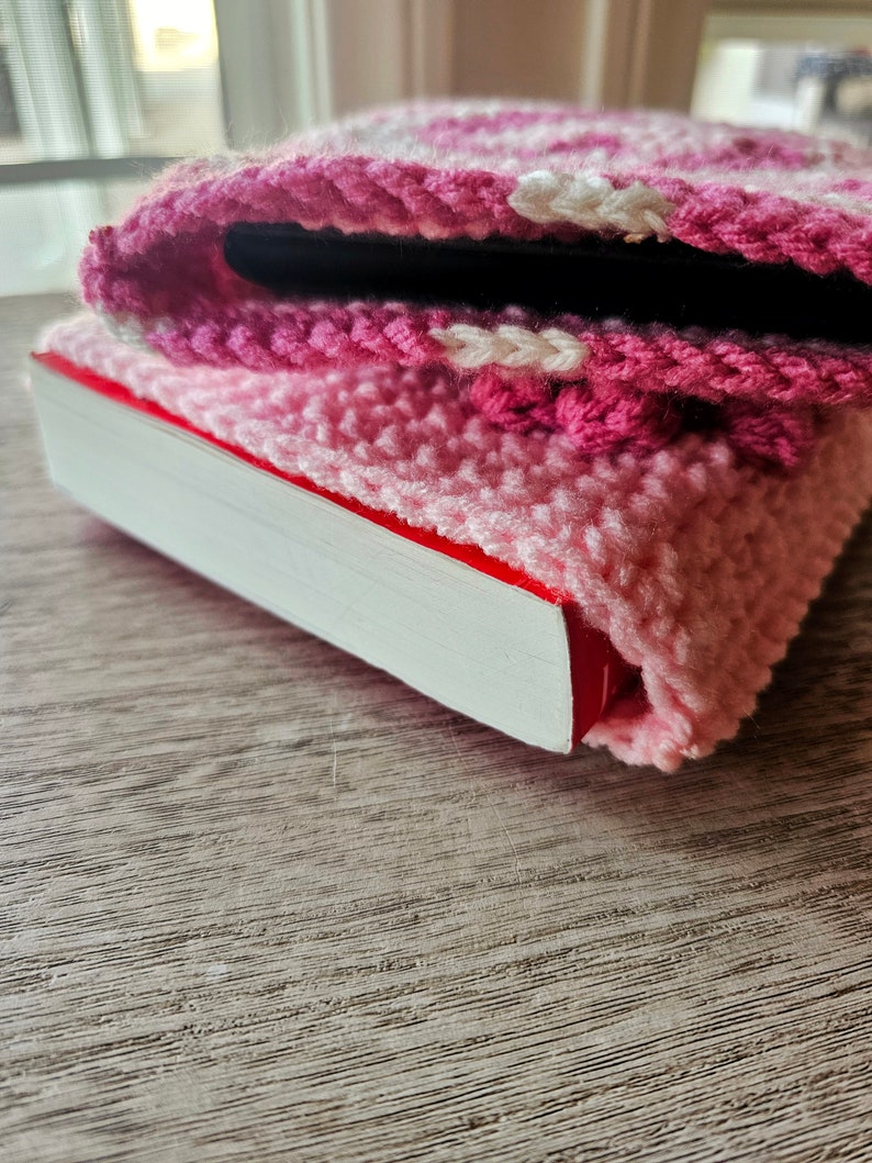 Puede incluir: Una funda de tel&eacute;fono de crochet rosa y blanca con una franja blanca, que cubre un libro con una encuadernaci&oacute;n roja.
