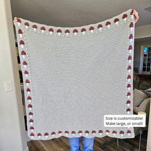 May include: A light gray crocheted blanket with a border of red and white mushroom designs. The blanket is held up, showing its rectangular shape. Text on the blanket reads: "Size is customizable! Make large, or small!"