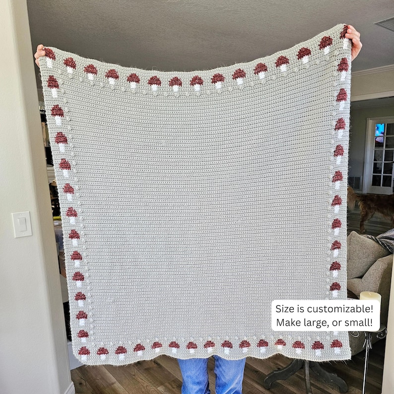 May include: A light gray crocheted blanket with a border of red and white mushroom designs. The blanket is held up, showing its rectangular shape. Text on the blanket reads: "Size is customizable! Make large, or small!"