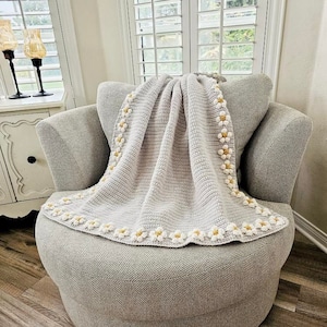 May include: A gray crocheted throw blanket with a white and yellow daisy border. The blanket is draped over a gray armchair.