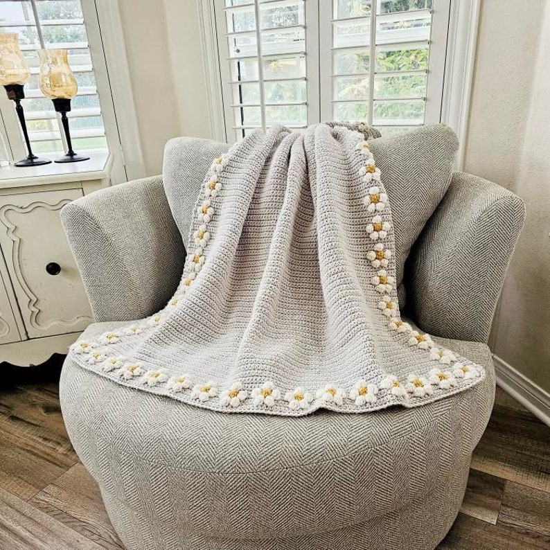 May include: A gray crocheted throw blanket with a white and yellow daisy border. The blanket is draped over a gray armchair.