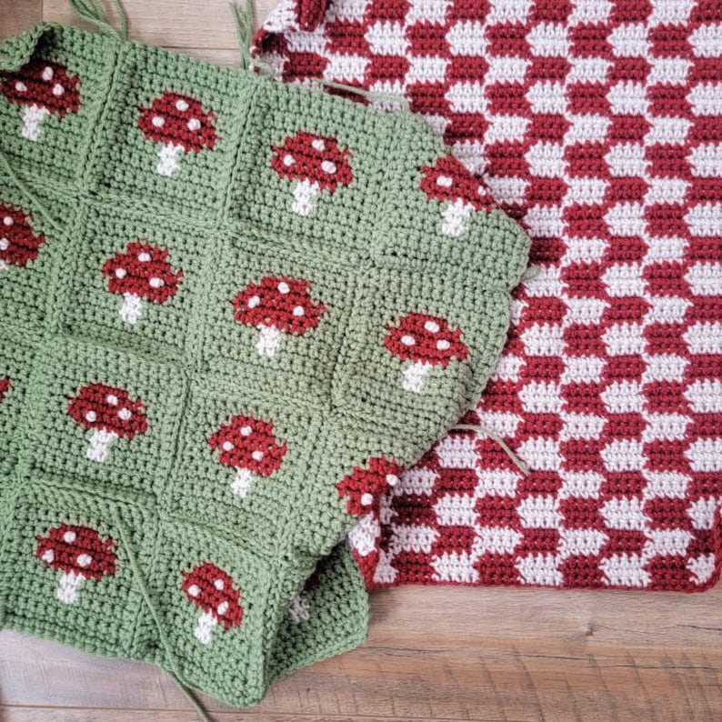 Puede incluir: Dos mantas de crochet, una verde con dise&ntilde;os de setas rojas y otra a cuadros rojos y blancos. Las mantas est&aacute;n dispuestas sobre una superficie de madera.