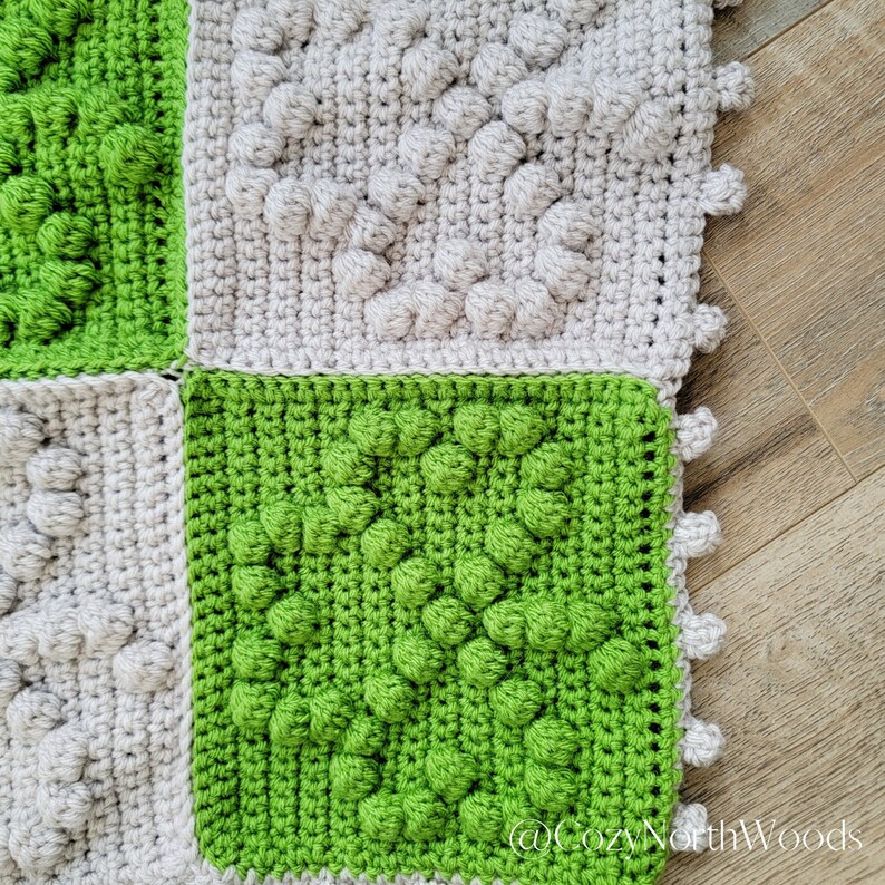 May include: Close-up of a crocheted blanket with squares in shades of green and gray. The squares have a textured, puffy design.