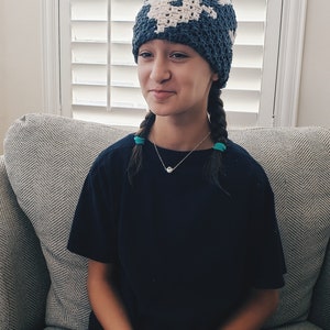 May include: A young person wearing a blue and white crocheted hat with a small orange pom-pom on top. The hat has a geometric pattern. The person is sitting on a gray couch and wearing a black t-shirt and dark shorts.