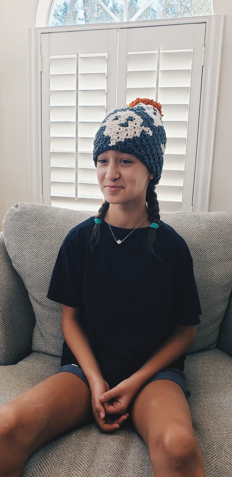 May include: A young person wearing a blue and white crocheted hat with a small orange pom-pom on top. The hat has a geometric pattern. The person is sitting on a gray couch and wearing a black t-shirt and dark shorts.