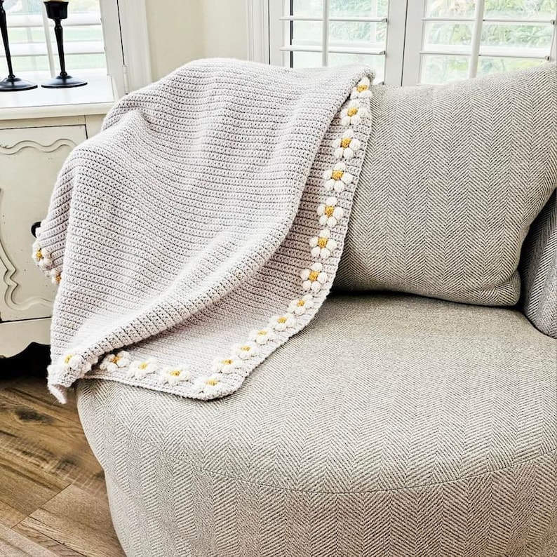 May include: A crocheted throw blanket with a white and yellow daisy trim. The blanket is draped over a gray armchair.