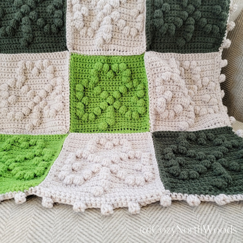 May include: A crocheted blanket with a patchwork design featuring squares in shades of green and beige. The squares have a textured, raised pattern.