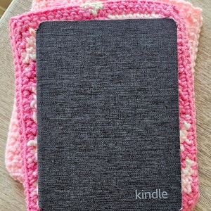 Puede incluir: Un lector electr&oacute;nico Kindle negro con la palabra "kindle" impresa en la parte delantera, descansando sobre un cuadrado de crochet rosa.