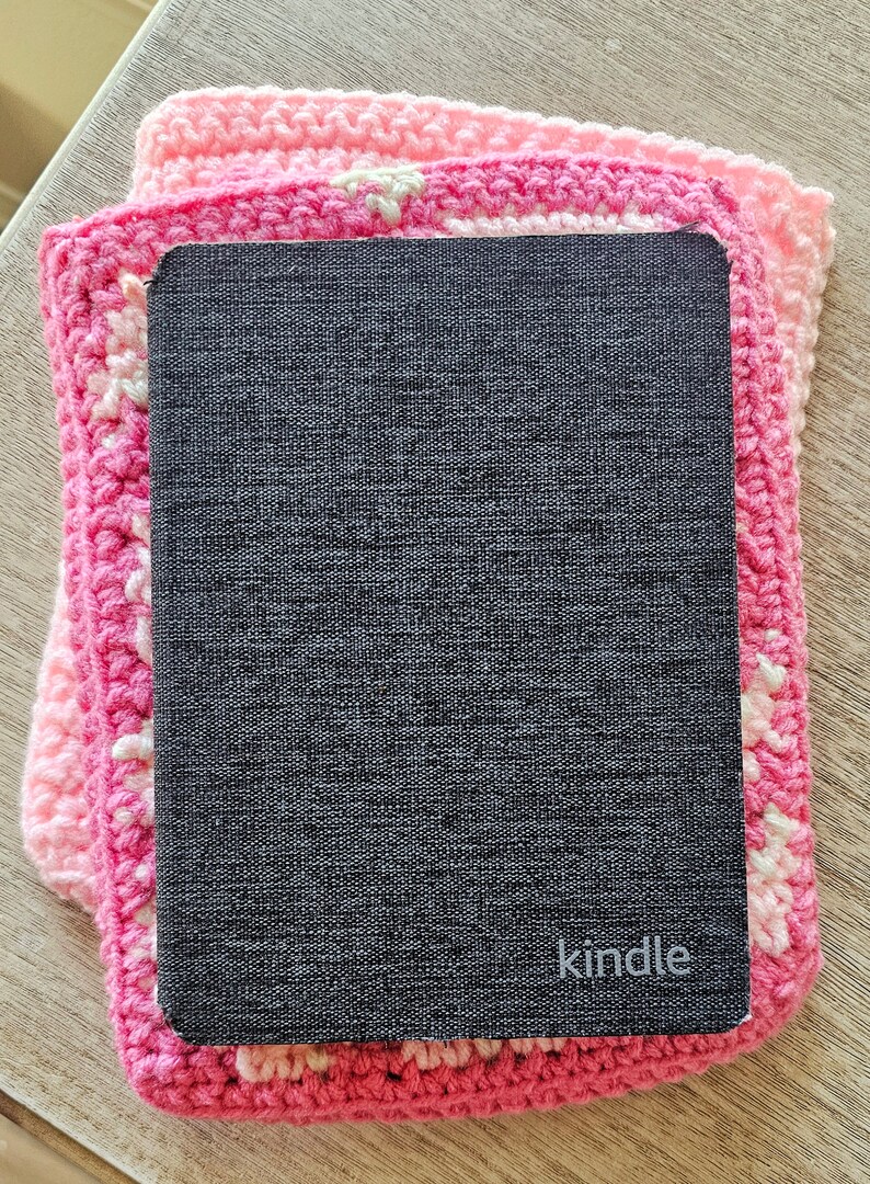 Puede incluir: Un lector electr&oacute;nico Kindle negro con la palabra "kindle" impresa en la parte delantera, descansando sobre un cuadrado de crochet rosa.