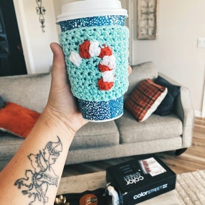 May include: A teal blue crocheted coffee cup cozy with a white and red candy cane design.