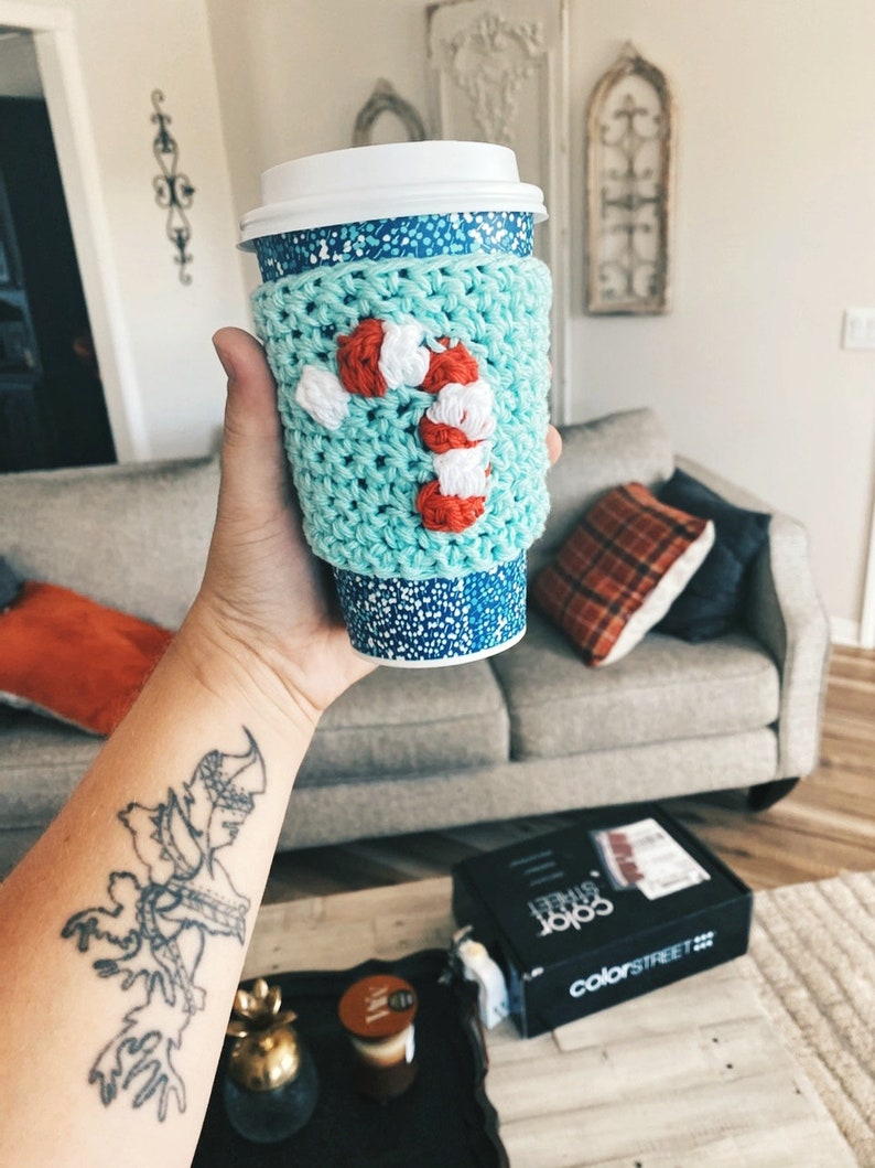 May include: A teal blue crocheted coffee cup cozy with a white and red candy cane design.