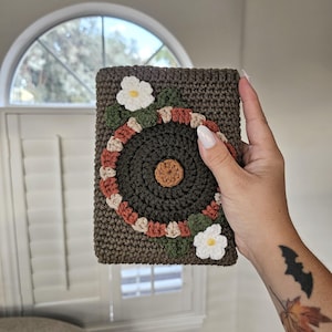 Puede incluir: Una bolsa de color marrón hecha a crochet con un diseño circular en tonos marrones, naranjas y blancos. La bolsa está decorada con flores blancas de crochet.