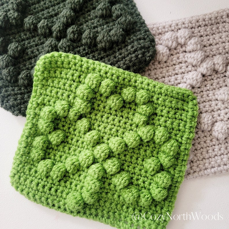 May include: Three crocheted squares in different shades of green and beige. The squares have a textured, bumpy surface.