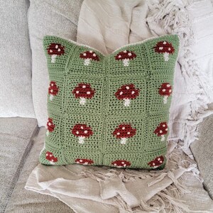 Puede incluir: Una almohada de crochet verde con un patr&oacute;n de hongos. La almohada est&aacute; hecha de un hilo suave y texturizado y tiene un borde blanco.