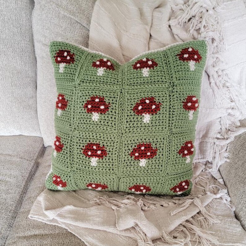 Puede incluir: Una almohada de crochet verde con un patr&oacute;n de hongos. La almohada est&aacute; hecha de un hilo suave y texturizado y tiene un borde blanco.