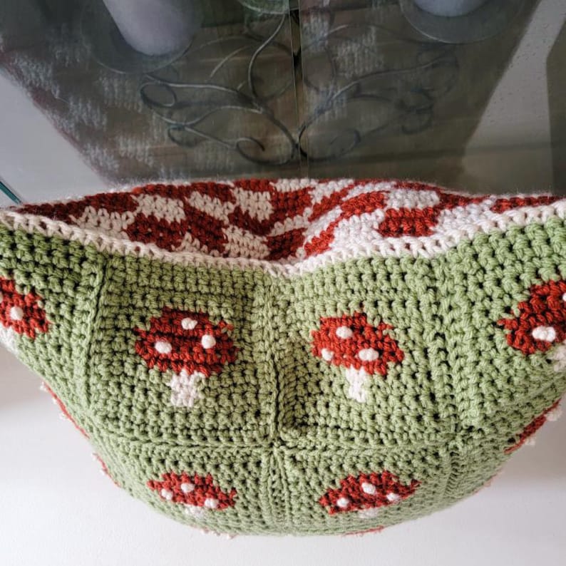 Puede incluir: Una almohada tejida a crochet con un patr&oacute;n de cuadros verdes y blancos y setas rojas. La almohada tiene forma de cuenco y una superficie suave y texturizada.