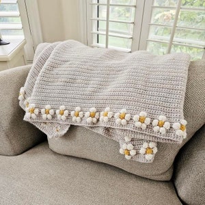 May include: A crocheted throw blanket in a light gray color with a border of white crocheted daisies with yellow centers. The blanket is draped over a gray couch.