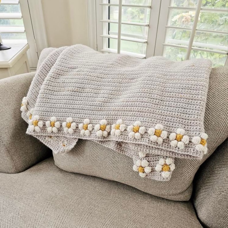 May include: A crocheted throw blanket in a light gray color with a border of white crocheted daisies with yellow centers. The blanket is draped over a gray couch.