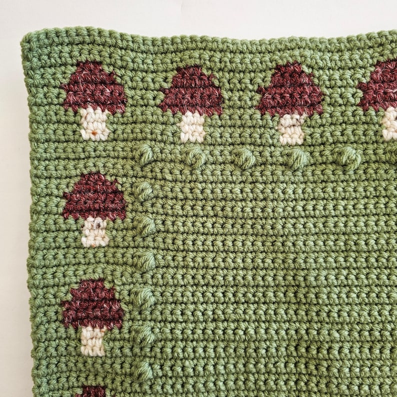 May include: A close-up of a crocheted green pillow cover with a repeating pattern of burgundy and cream-colored mushrooms. The mushrooms are evenly spaced across the surface, with textured, raised details. The overall aesthetic is handmade and whimsical.