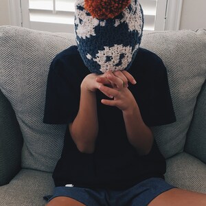 May include: A person wearing a blue and white crocheted hat with an orange pom-pom on top. The hat is covering the person's face. The person is sitting on a gray couch.