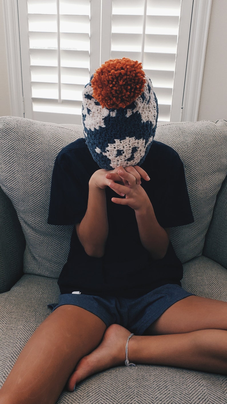 May include: A person wearing a blue and white crocheted hat with an orange pom-pom on top. The hat is covering the person's face. The person is sitting on a gray couch.