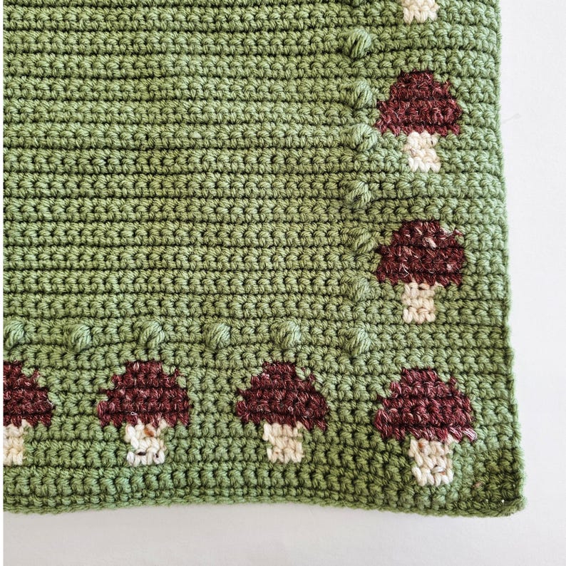 May include: A green crocheted blanket with a brown and white mushroom border. The blanket is made with a single crochet stitch and has a textured surface. The mushrooms are crocheted in a simple design with a brown cap and a white stem.