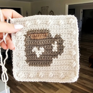 May include: A crocheted square with a brown and white design of a jar with a lid. The square is surrounded by a white border with raised bumps.