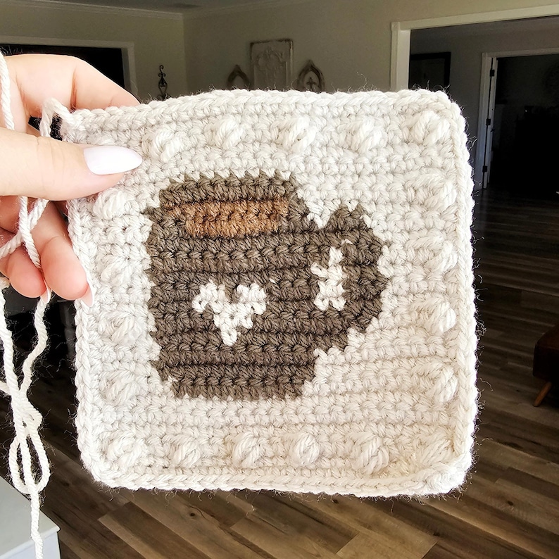 May include: A crocheted square with a brown and white design of a jar with a lid. The square is surrounded by a white border with raised bumps.
