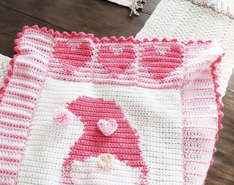 Puede incluir: Una manta de bebé de crochet rosa y blanca con un diseño de corazón y un gnomo rosa en el centro. La manta tiene un borde festoneado.