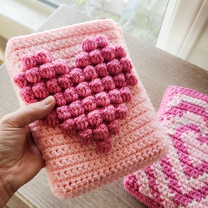 Puede incluir: Dos bolsas de coraz&oacute;n de punto de crochet rosa. Una bolsa tiene un dise&ntilde;o de coraz&oacute;n en relieve y la otra tiene un patr&oacute;n a rayas.
