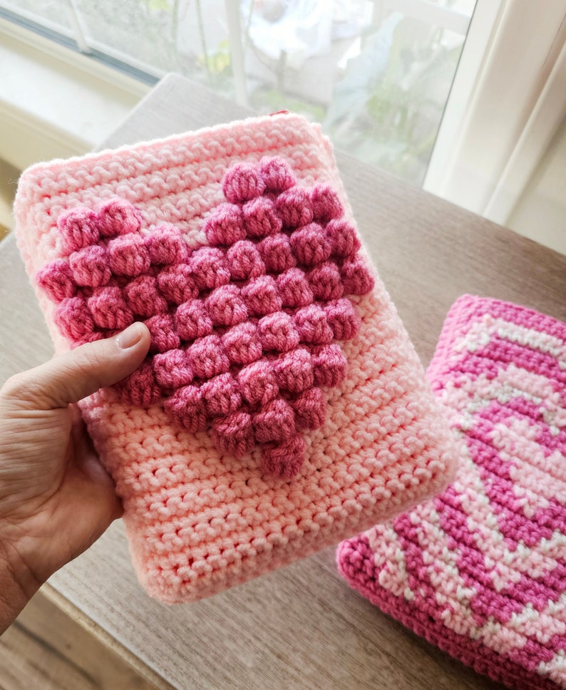 Puede incluir: Dos bolsas de coraz&oacute;n de punto de crochet rosa. Una bolsa tiene un dise&ntilde;o de coraz&oacute;n en relieve y la otra tiene un patr&oacute;n a rayas.