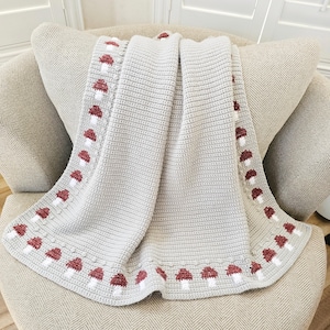 May include: A light gray crocheted blanket with a repeating pattern of red and white mushrooms along the border. The blanket is draped over a beige armchair, showcasing its texture and design. The blanket is made of soft material.