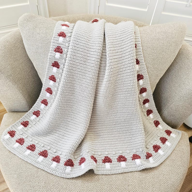 May include: A light gray crocheted blanket with a repeating pattern of red and white mushrooms along the border. The blanket is draped over a beige armchair, showcasing its texture and design. The blanket is made of soft material.