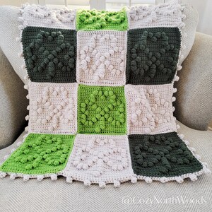 May include: A crocheted blanket with a checkered pattern in shades of green and beige. The blanket is made with a textured stitch that creates a raised, puffy effect.