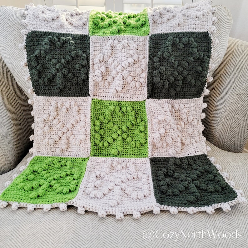May include: A crocheted blanket with a checkered pattern in shades of green and beige. The blanket is made with a textured stitch that creates a raised, puffy effect.