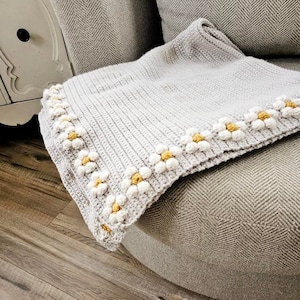 May include: A crocheted throw blanket with a gray background and a white daisy border with yellow centers. The blanket is draped over a gray armchair.