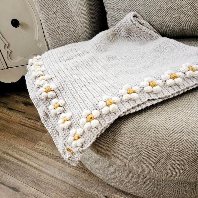 May include: A crocheted throw blanket with a gray background and a white daisy border with yellow centers. The blanket is draped over a gray armchair.