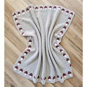May include: A light gray crocheted blanket with a repeating pattern of burgundy and white mushrooms along the border. The blanket is folded and draped, showcasing the texture and design. The blanket is made of soft yarn.