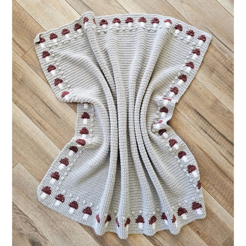 May include: A light gray crocheted blanket with a repeating pattern of burgundy and white mushrooms along the border. The blanket is folded and draped, showcasing the texture and design. The blanket is made of soft yarn.