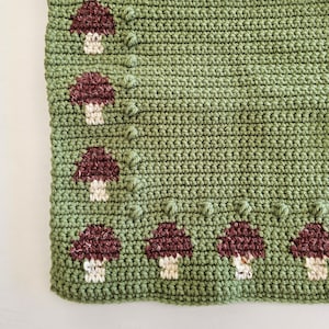 May include: A crocheted blanket in shades of green, featuring a repeating pattern of burgundy and cream-colored mushrooms. The blanket has a textured appearance due to the crochet stitches and mushroom details. The mushrooms are evenly spaced along the edge.