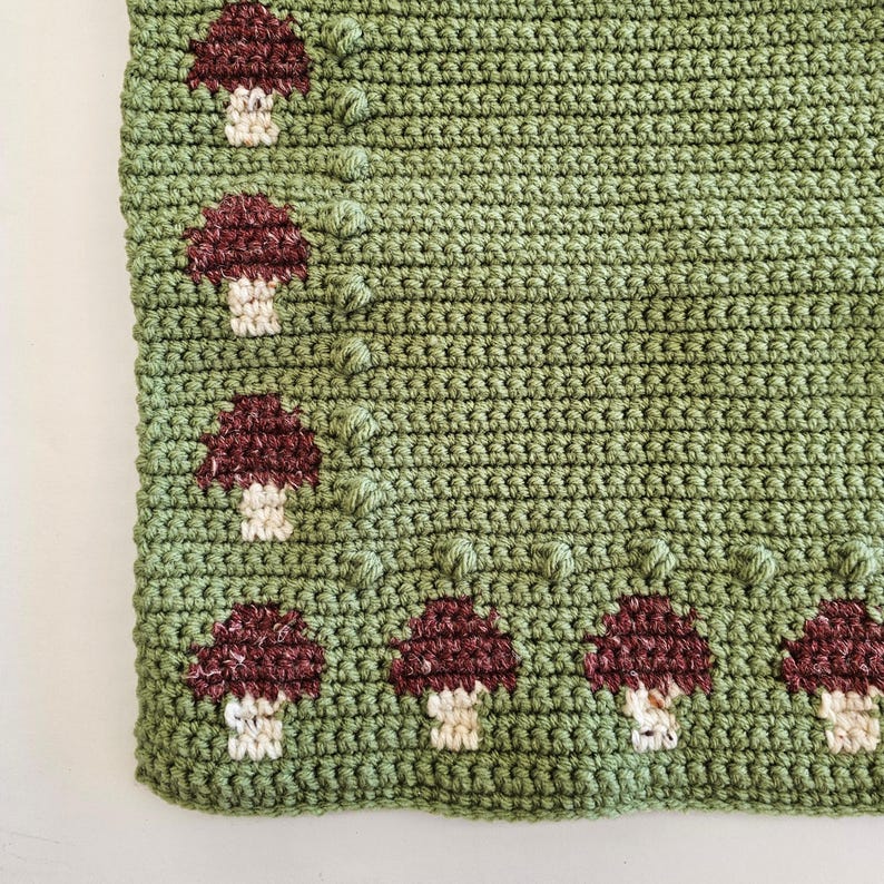 May include: A crocheted blanket in shades of green, featuring a repeating pattern of burgundy and cream-colored mushrooms. The blanket has a textured appearance due to the crochet stitches and mushroom details. The mushrooms are evenly spaced along the edge.